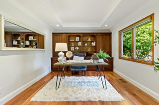 A cozy home office with a wooden desk, decorative items, and a lamp on a fluffy rug. Shelves with books and plants are behind, and a large window brightens the room.