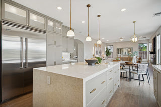 A_kitchen_featuring_light_gray_cabinetry_a_large_white_quartz_island_with_brass_hardware_stainless_steel_refrigerator_and_an_adjoining_dining_area.