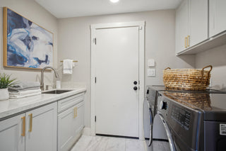A_laundry_room_featuring_light_gray_cabinetry_with_gold_hardware_a_white_countertop_sink_are_and_a_white_door_set_against_light_gray_walls.