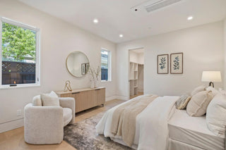 A modern bedroom with a cozy, neutral-toned aesthetic. It features a plush bed with layered pillows, a wooden dresser with a round mirror, and a soft armchair. Two windows allow natural light, illuminating plants in vases and botanical wall art.