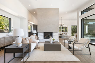 A modern entryway with a wooden door, textured gray console, white vase with branches, abstract wall art, and large window showing an outdoor patio .