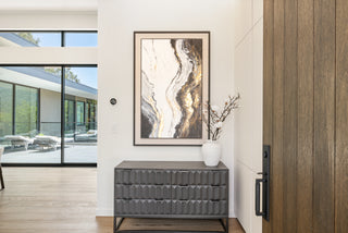 A modern entryway with a wooden door, textured gray console, white vase with branches, abstract wall art, and large window showing an outdoor patio.