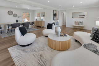 A modern living and dining area with wooden floors and white walls. Curved, white couches surround a round coffee table in the foreground. The dining area features a large table with six grey chairs. Accessories like cushions and lamps add a cozy touch. Natural light creates an inviting atmosphere.