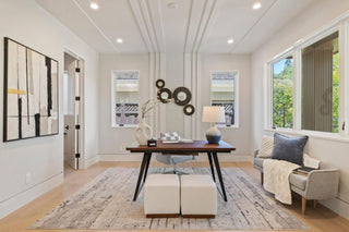 A modern, minimalist office with light walls and wooden flooring. Central desk with decor, abstract art on the wall, gray couch, and large windows. Cozy and bright