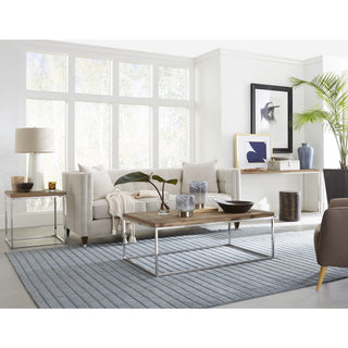 A reclaimed wood end table with a herringbone pattern tabletop and a stainless steel base, placed in a room with a sofa, coffee table, and armchair.