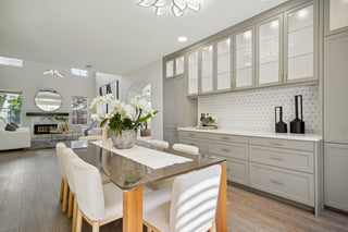 A_room_with_a_glass-top_table_six_upholstered_chairs_built-in_gray_cabinets_decorative_vases_and_a_view_into_a_living_area_with_a_fireplace_and_sofas