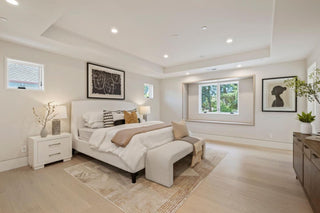 A spacious, modern bedroom with a large bed, neutral decor, and soft lighting. Light wood floors, artwork, and a window with greenery outside create a serene atmosphere.