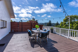 A spacious wooden deck with a dining table set and blue cushions under string lights. Surrounded by trees, it exudes a serene, inviting vibe.