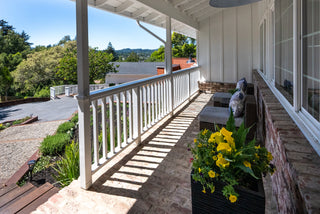 A sunny porch with white railings overlooking a garden. There’s a wicker chair with cushions and a planter of vibrant yellow flowers, creating a serene atmosphere.