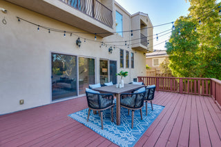 Backyard deck with a dining table and chairs on a blue rug, surrounded by trees and a house.