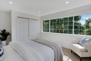 Bright bedroom with a large window showing green trees, a neatly made bed, beige armchair, blue pillow, plant, and white closet doors. Minimalist and serene.