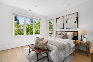 Bright bedroom with large windows showing greenery, featuring a cozy bed with pillows and a throw. An elegant bench, nightstand, and abstract art enhance the serene, modern vibe.