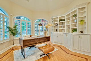Home office with wooden desk, white bookshelves, and large windows.