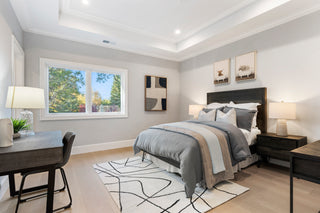 Bright, modern bedroom featuring a queen bed with gray bedding, light wood floors, a large window overlooking trees, and a dark wood desk area.