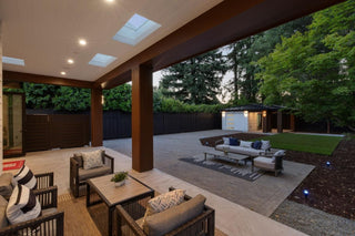Covered patio with plush outdoor seating, wooden pillars, and skylights. It overlooks a spacious yard with a modern shed, bordered by tall trees at dusk.