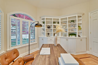 Home office with white bookshelves, leather chairs, and a large window.