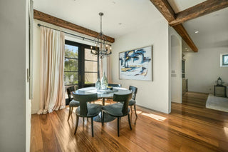 Dining room with wooden floors, a round glass table, and four black chairs. Modern art, a chandelier, and large windows create an inviting atmosphere.