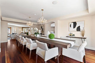 Elegant dining room with a long wooden table, white chairs, and two chandeliers. Decor includes green plants and abstract art, conveying a luxurious setting.