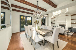 Elegant dining room with exposed wooden beams and a rustic table set for eight. A crystal chandelier adds a touch of luxury. Large windows and a fireplace create a cozy, inviting atmosphere.