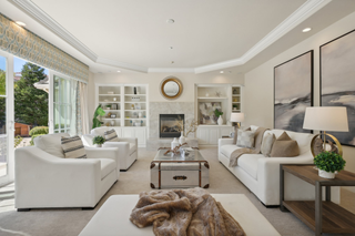 Elegant living room with white sofas, a glass coffee table, and decorative pillows. A fireplace, large windows, and abstract art create a cozy and sophisticated ambiance.