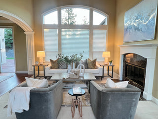 Elegant living room with white sofas, a glass coffee table, and decorative pillows. A fireplace, large windows, and abstract art create a cozy and sophisticateda ambiance.