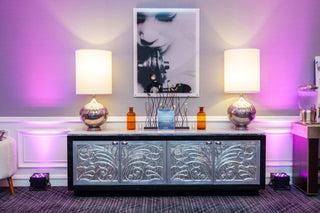 Decorative console table with ornate design, two lamps, and a framed picture in a room with purple lighting.