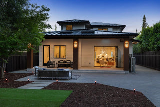 Latest house exterior at dusk with warm lights. Large glass doors open to reveal a stylish, inviting interior. Trees and a fence surround the patio.