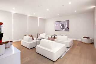 Minimalist living room with low white sofas, beige cushions, and a framed abstract painting on the wall. Soft lighting creates a calm atmosphere