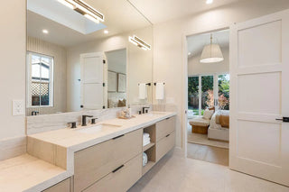 Modern bathroom with a large mirror and dual sinks. Light wood cabinetry and black fixtures. Open door reveals a cozy living room with sofa and bright lamp.