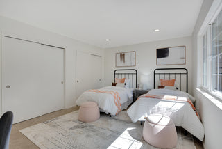 Modern bedroom with two single beds featuring white and orange bedding, black metal frames, and matching artwork. Soft lighting and neutral tones create a calm, inviting atmosphere. Pink poufs and a patterned rug complete the decor.
