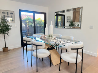 Modern dining room with a glass table and six white chairs on a light wood floor. A large mirror reflects the room, plants, and decor accents create a cozy feel.