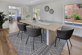 Modern dining room with a light wood table, six dark gray chairs on a geometric rug. Decor includes two vases and wall baskets, exuding elegance.