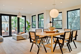 Modern dining room with a wooden table, six chairs, and a large round light fixture. Sunlight streams through large windows, highlighting a cozy, inviting atmosphere.