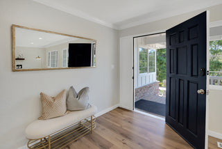 Modern entrance with a black door open to a porch. Inside, a bench with pillows sits below a wall mirror on a light gray wall. Bright, welcoming ambiance.