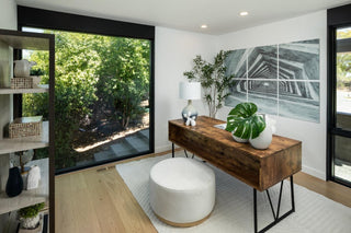 Modern home office with wooden desk, white ottoman, and large windows.
