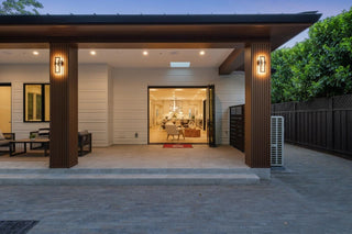 Modern house exterior at dusk with warm lights. Large glass doors open to reveal a stylish, inviting interior. Trees and a fence surround the patio.