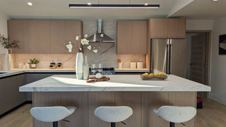 Modern kitchen with light wood cabinets and marble countertops. A large island features vases with flowers, lemons, and wine glasses. Warm and inviting.