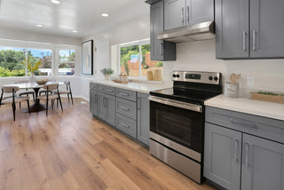 Modern kitchen with sleek gray cabinets, stainless steel stove, and wooden floors. Sunlit dining area with glass table and chairs by large windows.