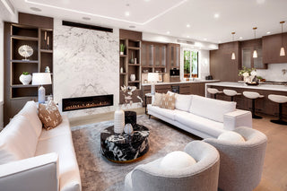 Modern living room and kitchen with white sofas and armchairs, marble fireplace, and wooden cabinetry. Soft lighting creates a cozy ambiance.