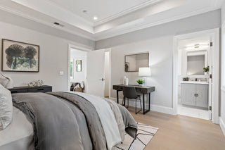 Modern master bedroom with light gray walls, light wood floors, a gray bedspread, a dark wood desk, and an ensuite bathroom visible through an open doorway.