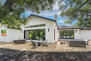 Modern one-story house with white exterior, large windows, and sloped roof. Outdoor seating and dining area under lush trees create a serene setting.