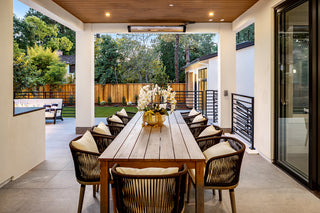 Modern outdoor dining area with a long wooden table, surrounded by cushioned chairs. Elegant flowers in a vase. Warm lighting sets a cozy, inviting tone.
