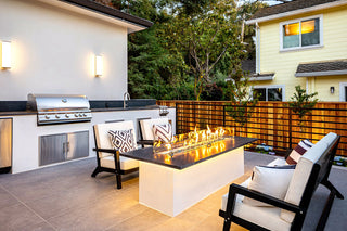 Modern patio with a sleek grill, cozy chairs with patterned cushions, and a long fire pit table. Warm lighting and wooden fencing create a welcoming ambiance.