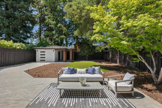 Modern patio with wicker seating under a covered roof with skylights. Overlooks a spacious backyard with a wooden fence and sofa, lush green trees, and a small shed.