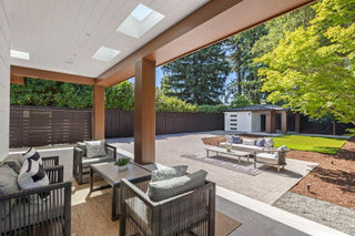 Modern patio with wicker seating under a covered roof with skylights. Overlooks a spacious backyard with a wooden fence, lush green trees, and a small shed.