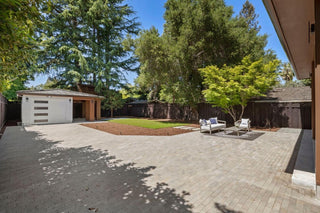 Modern patio with wicker seating under a covered roof with skylights. Overlooks a spacious backyard with a wooden fence, lush green trees, and a small shed.
