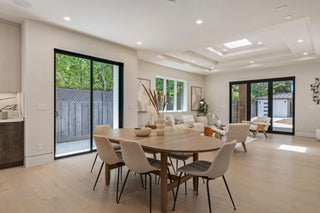 Modern, spacious living room with beige tones, featuring a wood dining table, gray chairs, plush sofas, large windows, and sliding glass doors. Bright, airy atmosphere.