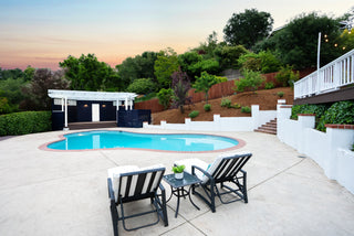 Pool area with lounge chairs and a table on a patio, surrounded by greenery.