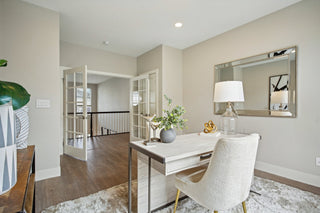 Bright_home_office_featuring_a_light_desk_cream_chair_glass_lamp_and_mirrored_wall_decor_opening_with_French_doors_overlooking_a_staircase.
