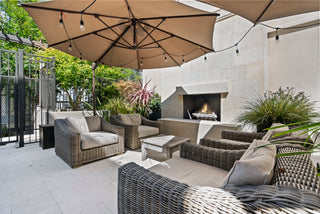Outdoor patio with wicker furniture under large umbrellas. A cozy fireplace is surrounded by lush greenery, creating a relaxing and inviting atmosphere.